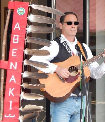 Bryan Blanchette in white shirt, black vest, faded blue jeans and wearing sun glasses, has his guitar over is shoulders and is playing, next to a long thin red flag that spells abenaki